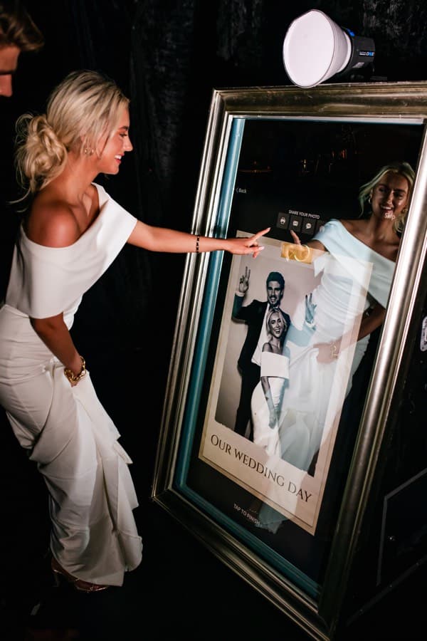 Bride tapping the sharing screen of a gilt-framed mirror photo booth at a wedding reception